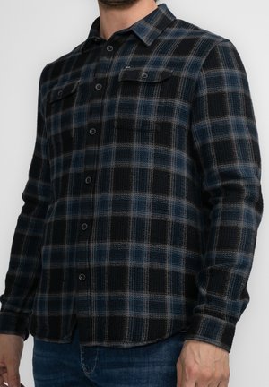 Homme portant une chemise à manches longues noire et bleue à carreaux avec deux poches poitrine et un jean bleu foncé.