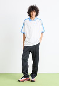 Camisa polo branca de manga curta com detalhes azuis, combinada com calças de desporto pretas e sapatilhas de corrida multicoloridas com um design de sola em bolha.