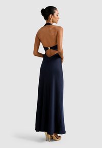 Mulher com o cabelo apanhado em um coque, vestindo um vestido halter longo, azul-marinho, com as costas abertas e sandálias de salto alto douradas, de pé de lado.