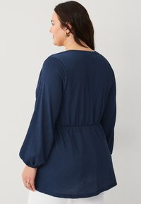 Navy blue long-sleeve blouse with a textured finish. Elastic waist detailing and gathered cuffs. Soft, flowing design with a round neckline.