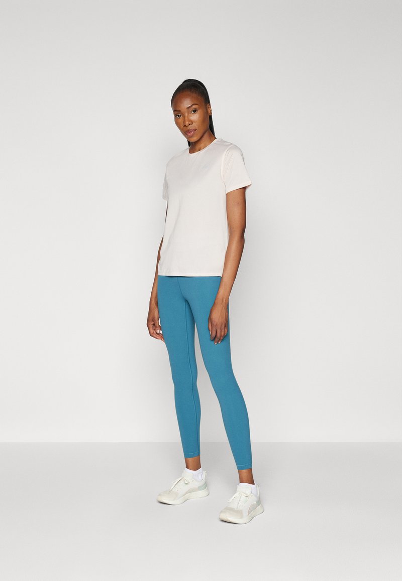 T-shirt rose clair à manches courtes associé à des leggings bleus et des chaussures de sport blanches. Le tissu semble lisse avec un design ajusté.