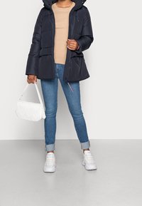 Mørkeblå pufferjakke med hætte, ribbet beige sweater, blå jeans, hvid quilted håndtaske og hvide sneakers. Glatte teksturer og lagdelt design.