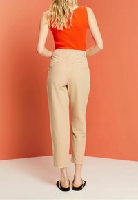Femme aux longs cheveux blonds porte un haut orange sans manches, un pantalon beige taille haute et des sandales noires, face à un mur orange.