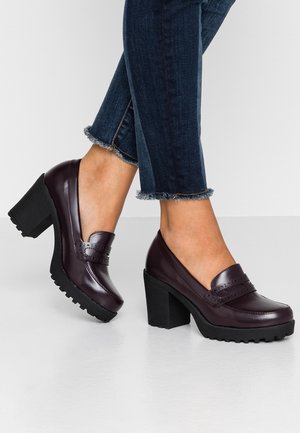 Frau trägt dunkelviolette Leder-Loafer mit klobigen Absätzen, profilierten Sohlen und Jeans mit ausgefranstem Saum.