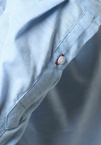 Țesătură de cămașă de culoare albastru deschis, amestec de bumbac. Prezintă un buton accent roșu și cusături detaliate aproape de guler și de manșetă. Textură fină.