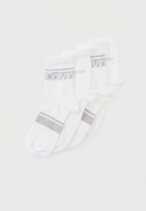 Četras baltas sporta zeķu pāri ar rievotu tekstūru, pelēkām purngala un papēža joslām, kā arī "Calvin Klein" logotipu uz aprocēm, sakārtotas pārklājoties.