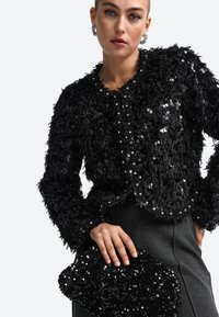 Musthave musta sädelev jakk, millel on tekstuuriline, pehme disain, ja sobitatud sädelev käekott. Mudel kannab halli seeliku.