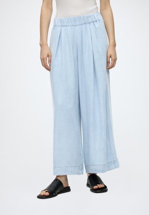 Persona que lleva pantalones anchos de color azul claro con cintura elástica y sandalias negras abiertas de tipo slide, de pie frente a un fondo blanco.