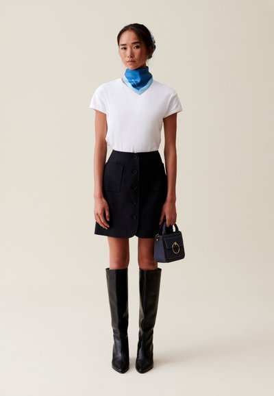 Top blanco de manga corta, falda negra en línea A con botones, botas de cuero negras hasta la rodilla y un pañuelo de seda azul; bolso pequeño azul con detalles en dorado.
