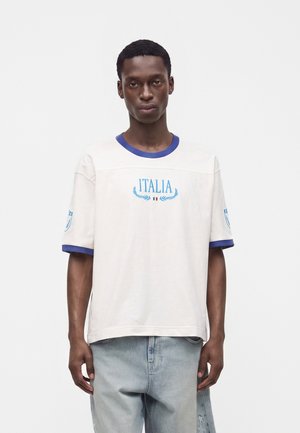 Giovane uomo che indossa una T-shirt bianca con rifiniture blu e testo "ITALIA", abbinata a jeans azzurri, in piedi su uno sfondo bianco semplice.