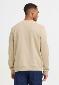 Felpa beige con scollo a girocollo e polsini a costine, abbinata a jeans blu scuro. Tessuto morbido, design semplice, senza loghi visibili.