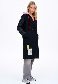 Žena stojí, má na sobě černý dlouhý kabát s růžovou kapucí, šedé džíny a bílé tenisky na bílém pozadí.