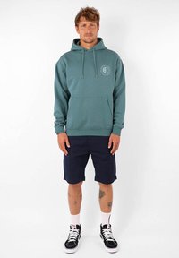 Hoodie teal avec une poche avant, des cordons ajustables et une impression de logo. Associé à un short bleu marine et des baskets noires. Chaussettes blanches à mi-mollet.