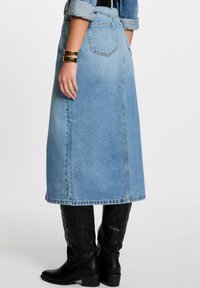 Lichte blauwe denim midi rok met een split aan de achterkant en opgestikte zakken. Gecombineerd met zwarte leren laarzen met puntige neuzen, wat de textuur benadrukt.