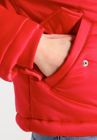Czerwona pikowana kurtka z kieszenią zapinaną na zatrzask, ściągaczami na rękawach oraz gładkim, błyszczącym wykończeniem.