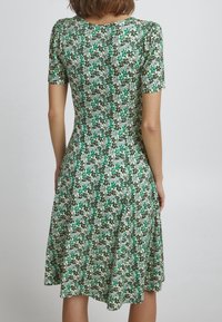 Robe florale verte avec des manches courtes, corsage cintré et jupe évasée. Elle est réalisée dans un tissu léger et doux, avec un motif de petites fleurs et feuilles.