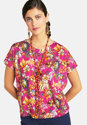 Woman with dark hair wearing a pink floral blouse, long red beaded necklace, colorful dangling earrings, and dark blue pants.