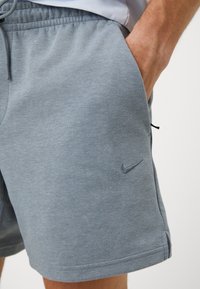 Gros plan d'une personne portant un short Nike gris avec une main dans la poche, sur un fond clair uni.