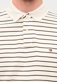 Μπεζ πόλο μπλουζάκι με λεπτές καφέ ρίγες, ριμπ κολάρο, πατιλέτα με τρία κουμπιά και μικρό κεντημένο λογότυπο στο αριστερό στήθος.