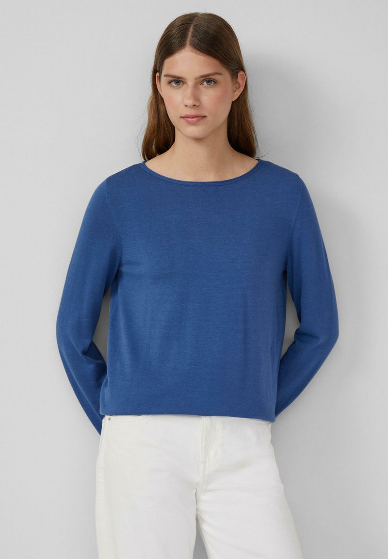 Langärmliges, blaues Strickoberteil mit rundem Ausschnitt und lässiger Passform, kombiniert mit weißen Hosen. Weiche Textur, minimalistisches Design.
