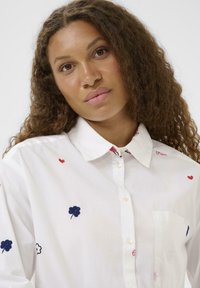 Egy göndör barna hajú nő fehér gombos inget visel, amelyen kicsi hímzett piros szívek és kék virágok láthatók, egy sima háttér előtt.