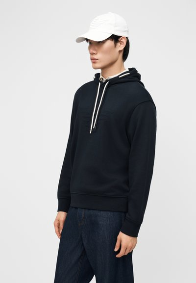 Νέος άνδρας που φοράει μαύρο φούτερ με λευκά κορδόνια, σκούρο τζιν και λευκό καπέλο μπέιζμπολ, κοιτάζοντας προς τα αριστερά μπροστά σε απλό φόντο.