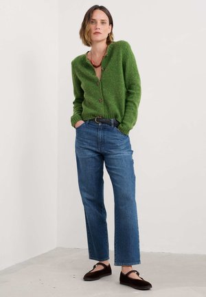 Donna che indossa un cardigan verde con bottoni, jeans blu a vita alta con cintura nera, scarpe flat marroni e una collana rossa, in piedi con le mani nelle tasche.