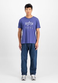 Lilla puuvillane t-särk, millel on valge "ALPHA INDUSTRIES" logo, koos lõtvade siniste teksade ja hallide spordijalatsitega. Lihtne disain.