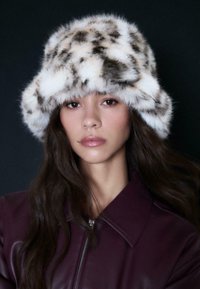 Una donna con lunghi capelli scuri indossa un cappello a secchiello in finta pelliccia bianca e marrone maculata e una giacca di pelle viola scuro su uno sfondo nero.