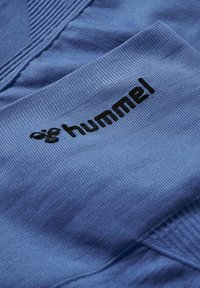 Blauer strukturierter Stoff mit schwarzem, besticktem "hummel"-Logo und Bienen-Symbol am Ärmel oder Bund eines Kleidungsstücks.