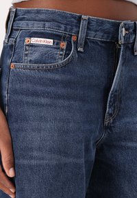 Tmavě modré džíny z denimu s hladkou texturou, na zadní kapse s označením značky Calvin Klein a železnými doplňky v barvě mědi.