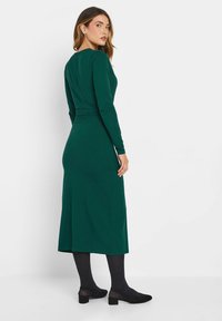 Grünes, langärmliges Kleid mit taillierter Passform und fließendem Rock, aus dehnbarem Stoff. Stylisch kombiniert mit schwarzen Strumpfhosen und Schuhen. Einfaches Design.