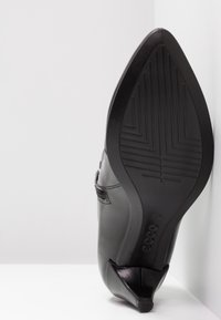 Chaussure à talon en cuir noir positionnée semelle vers le haut contre un coin blanc, montrant un bout pointu et une semelle en caoutchouc texturée avec le nom de la marque « ecco ».