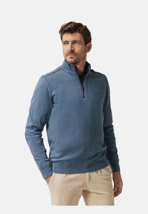 Blauer Strickpullover mit halbhohem Reißverschlusskragen, strukturiertem Stoff, langen Ärmeln und gerippten Bündchen, kombiniert mit hellen Hosen.