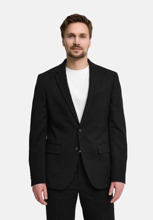 Mężczyzna z krótkimi brązowymi włosami i brodą, ubrany w czarną marynarkę i spodnie na białej koszuli, stojący na białym tle.