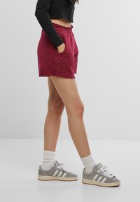 Burgunderfargede shorts med en frynset kant, elastisk liv, og sidelommer, med brodert design. Kombinert med grå joggesko og hvite sokker.