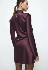 Vestido de satén burdeos con mangas largas, cuello alto y detalle de abertura en la espalda. Presenta una textura suave y una silueta ajustada.