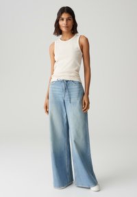 Top senza maniche beige chiaro abbinato a jeans di denim azzurro a gamba larga. I jeans presentano una vita con cordoncino e una texture di tessuto sbiadita.