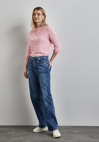 Helles rosa strukturiertes Sweatshirt mit dreiviertel Ärmeln, kombiniert mit hellblauen weit geschnittenen Jeans und weißen Turnschuhen, vor einem neutralen Hintergrund stehend.