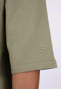 T-shirt in cotone verde oliva con maniche corte e una piccola lettera "B" ricamata sul polsino. Tessuto morbido e vestibilità casual.