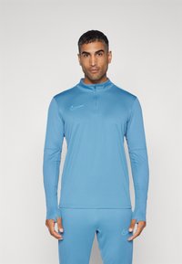 Világoskék Nike hosszú ujjú sport póló, fél cipzárral, testhezálló ujjakra és egy kontrasztos színű hímzett logóra.