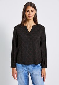 Kvinna med långt brunt hår iförd en svart mönstrad långärmad blus och ljusblå jeans som står mot en neutral bakgrund.