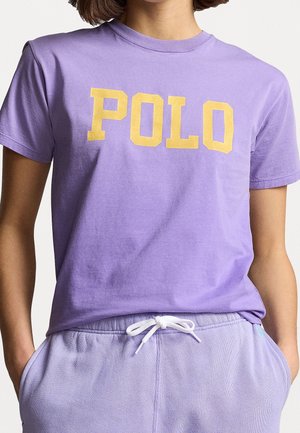 Henkilö pukeutuneena laventelinväriseen t-paitaan, jossa isolla keltaisella lukee "POLO", ja laventelinvärisiin vetokärpäsnauhalla varustettuihin housuihin, kädet taskuissa.