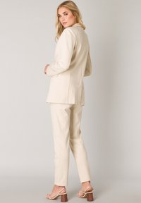 Crème blazer en bijpassende broek, op maat gemaakt, gladde textuur, minimale naden, met een enkele achterkant splits en subtiele stikdetails.