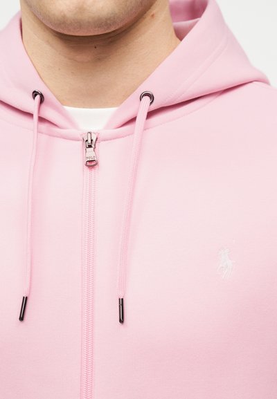 Άνδρας που φοράει ανοιχτό ροζ φούτερ με φερμουάρ και κορδόνια, με μικρό κεντημένο λογότυπο στο στήθος.