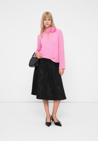 Klarr rosa långärmad blus med en blommig dekoration vid halsen, kombinerad med en texturerad svart midikjol och svarta spetsiga klackar.