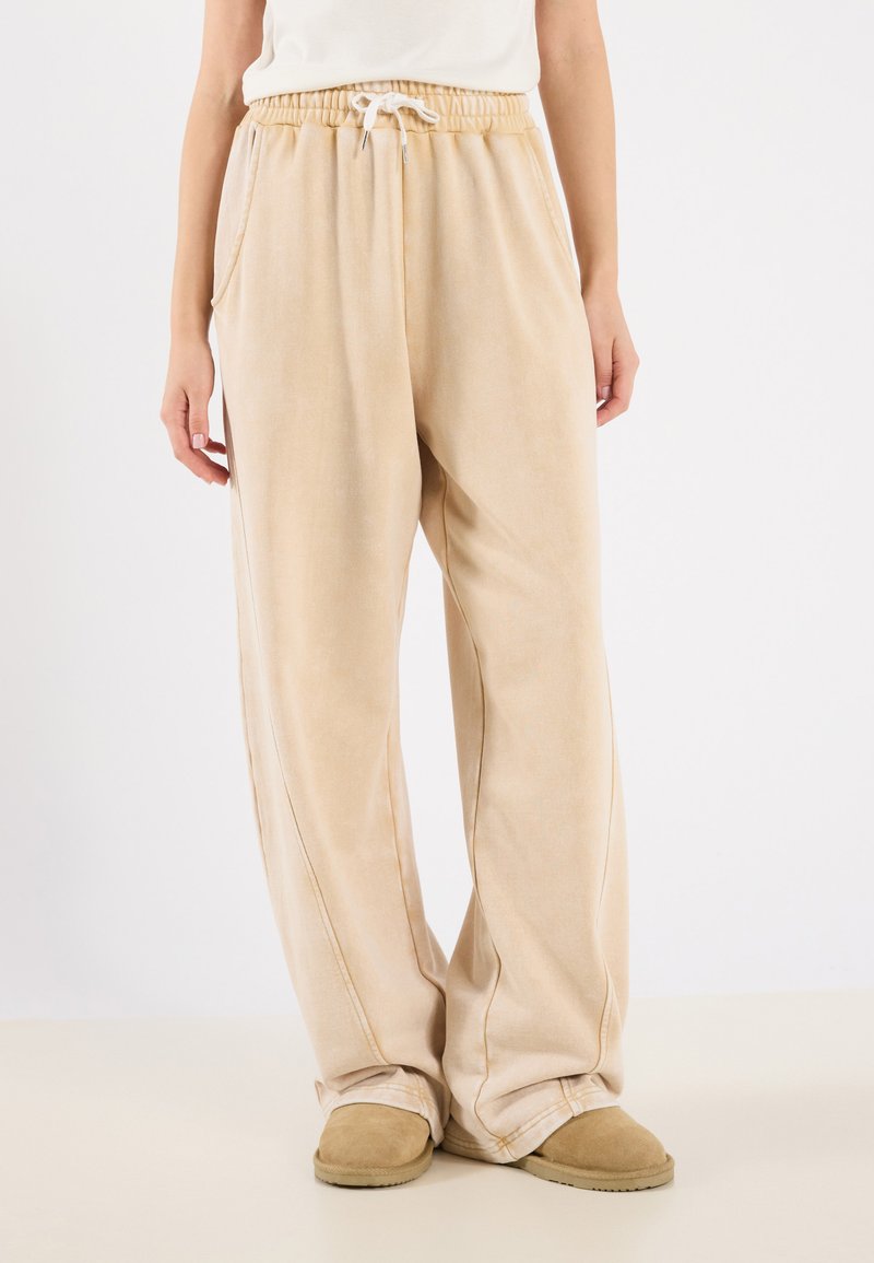 Beige joggebukser laget av myk stoff, med elastisk midje med snor, side lommer og kontrasterende sømdetaljer.