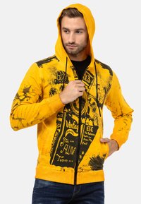 Sweat à capuche zippé jaune avec des designs graphiques noirs, un capuchon et un tissu texturé. Les détails notables incluent un imprimé vintage et des poches avant.