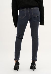 Vaqueros ajustados de denim azul oscuro, con una cintura de altura media, bolsillos traseros sin costuras y un ligero desgaste. Combinados con zapatos de tacón negro.
