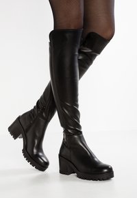 Las botas negras de cuero sobre la rodilla cuentan con un tacón grueso, suela texturizada, cremallera lateral y un acabado liso con detalles mínimos de costura.